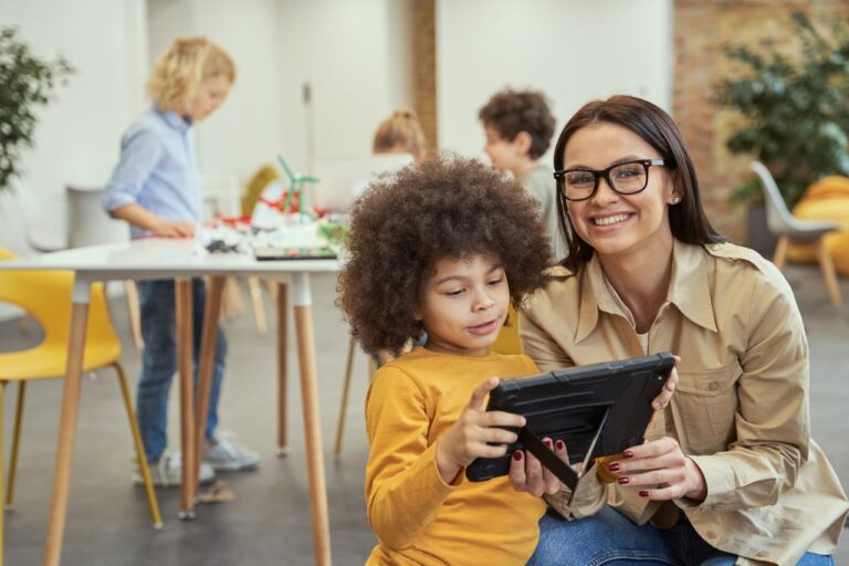 portrait-of-nice-young-female-teacher-smiling-at-camera-while-showing-video-on-tablet-pc-to-her.jpg